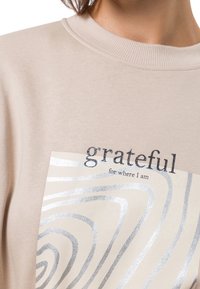 Beige katoenen sweatshirt met een zilveren folieafdruk van golvende lijnen en de tekst "grateful for where I am" in een contrasterend lettertype.
