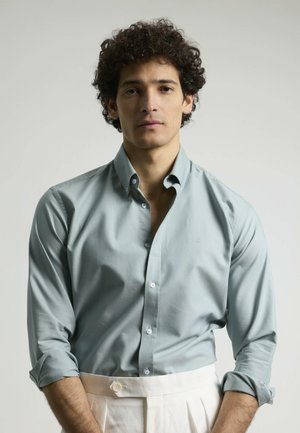 Camisa de botones de color azul claro con mangas remangadas, fabricada en un tejido suave. Presenta un corte ajustado y un sutil bordado de logo cerca del pecho.