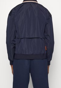 Person som bär en marinblå jacka med fackdetalj på ryggen och matchande marinblå byxor, står mot en enkel vit bakgrund.