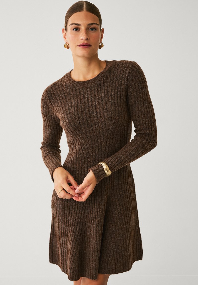 Frau in einem langen braunen Rippstrickkleid mit langen Ärmeln, goldenem Ohrschmuck und einem goldenen Armband, steht vor einem schlichten Hintergrund.