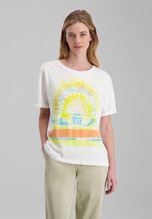 Young woman with light hair wears a white t-shirt with a colorful sunburst design and word "Libertad," paired with light beige pants.