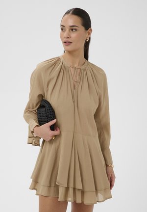 Femme en robe longue beige à manches longues superposées tenant une pochette noire tissée, portant des boucles d'oreilles dorées et un bracelet, regardant sur le côté.