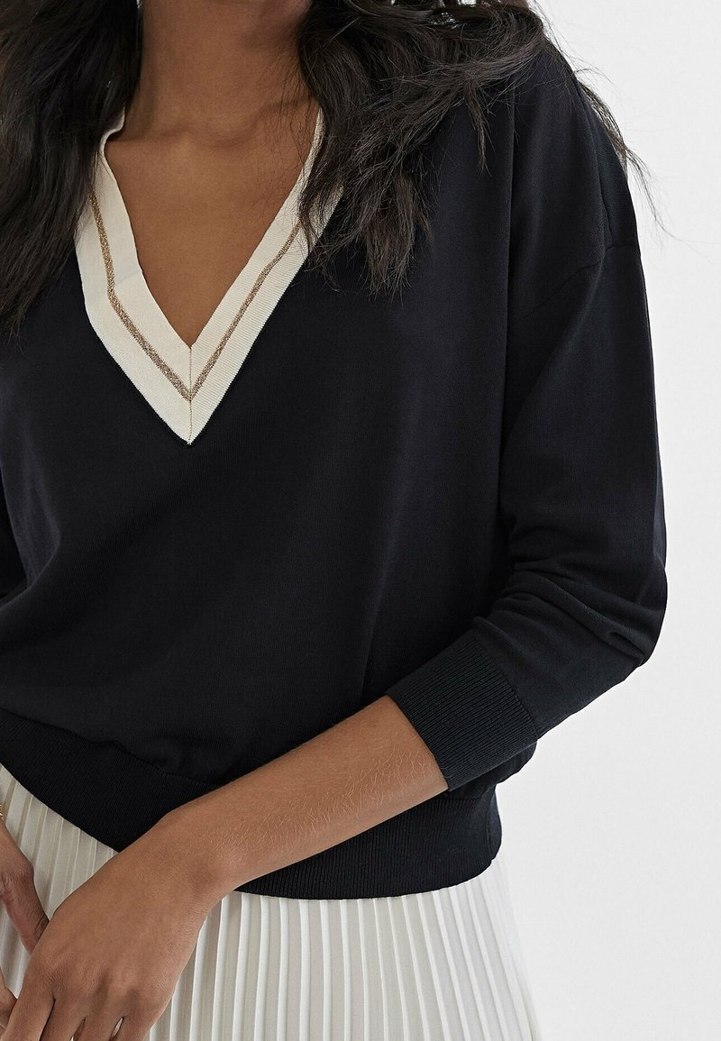 Un pull noir à col en V avec une bordure crème et dorée métallique, doté de manches longues et d'un ourlet côtelé, associé à une jupe blanche plissée.