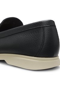 Gros plan du talon et du côté d’une chaussure en cuir noir texturé avec semelle en caoutchouc blanche sur fond blanc.