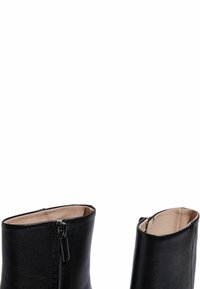 Botas de cuero negro hasta el tobillo con una textura suave, que cuentan con una cremallera lateral y un diseño de parte superior abierta. El interior está forrado con un color claro.