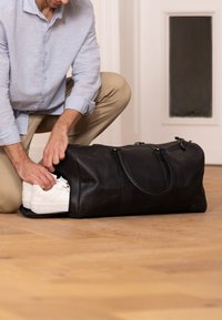 Mustassa nahkaisessa duffel-laukussa, joka on suuri ja tukevilla kahvoilla varustettu, pakataan valkoisia lenkkareita. Puulattia ja neutraali tausta korostavat laukun huomiota.