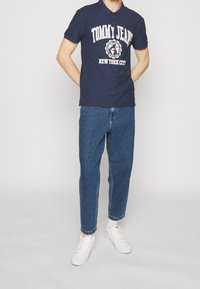 Man som bär en marinblå polotröja med vit "Tommy Jeans New York City"-logotyp, blå jeans, vita Nike-sneakers, står med händerna bakom ryggen.