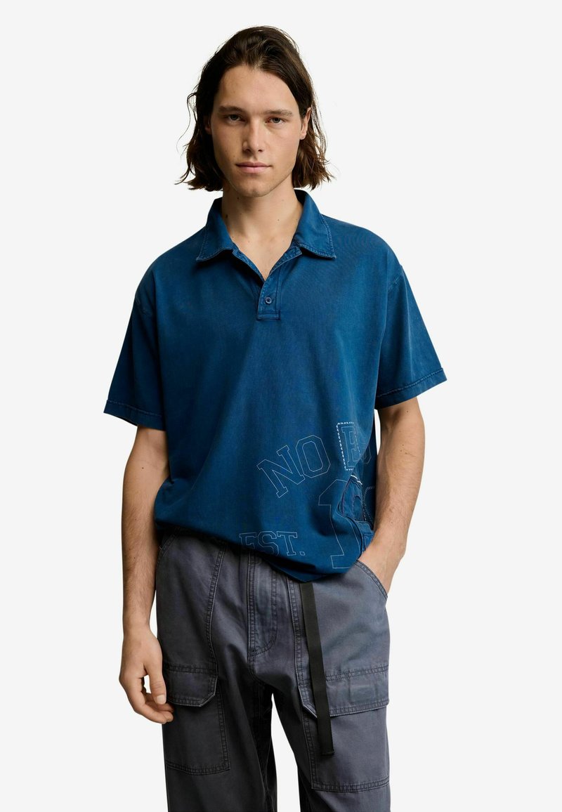 Jeune homme aux cheveux mi-longs portant un polo bleu ample et un pantalon cargo gris, debout avec une main dans la poche.