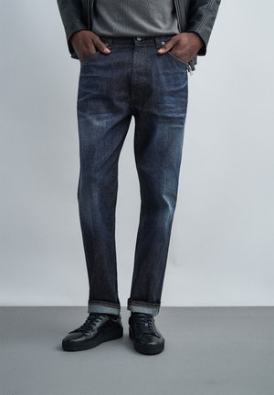 Personne portant un jean bleu foncé retroussé, des baskets noires, une chemise grise et une veste en cuir noire les mains dans les poches, debout sur un sol gris.