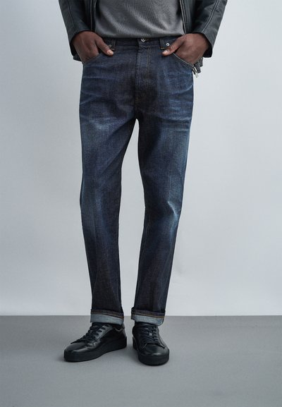 Person wearing dark blue cuffed jeans, black sneakers, gray shirt, and black leather jacket with hands in pockets, standing on gray floor.