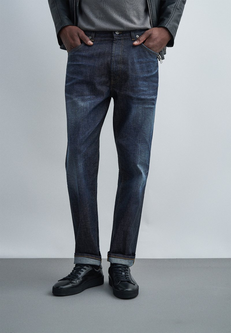 Άτομο που φοράει σκούρα μπλε τζιν με γυρισμένα ρεβέρ, μαύρα αθλητικά, γκρι πουκάμισο και μαύρο δερμάτινο μπουφάν με τα χέρια στις τσέπες, στέκεται σε γκρι πάτωμα.