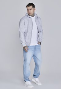 Szara bluza z zamkiem na zamek błyskawiczny, noszona na białej koszulce, w połączeniu z jasnoniebieskimi dżinsami i białymi trampkami. Bluza ma sznurek do regulacji i boczne kieszenie.