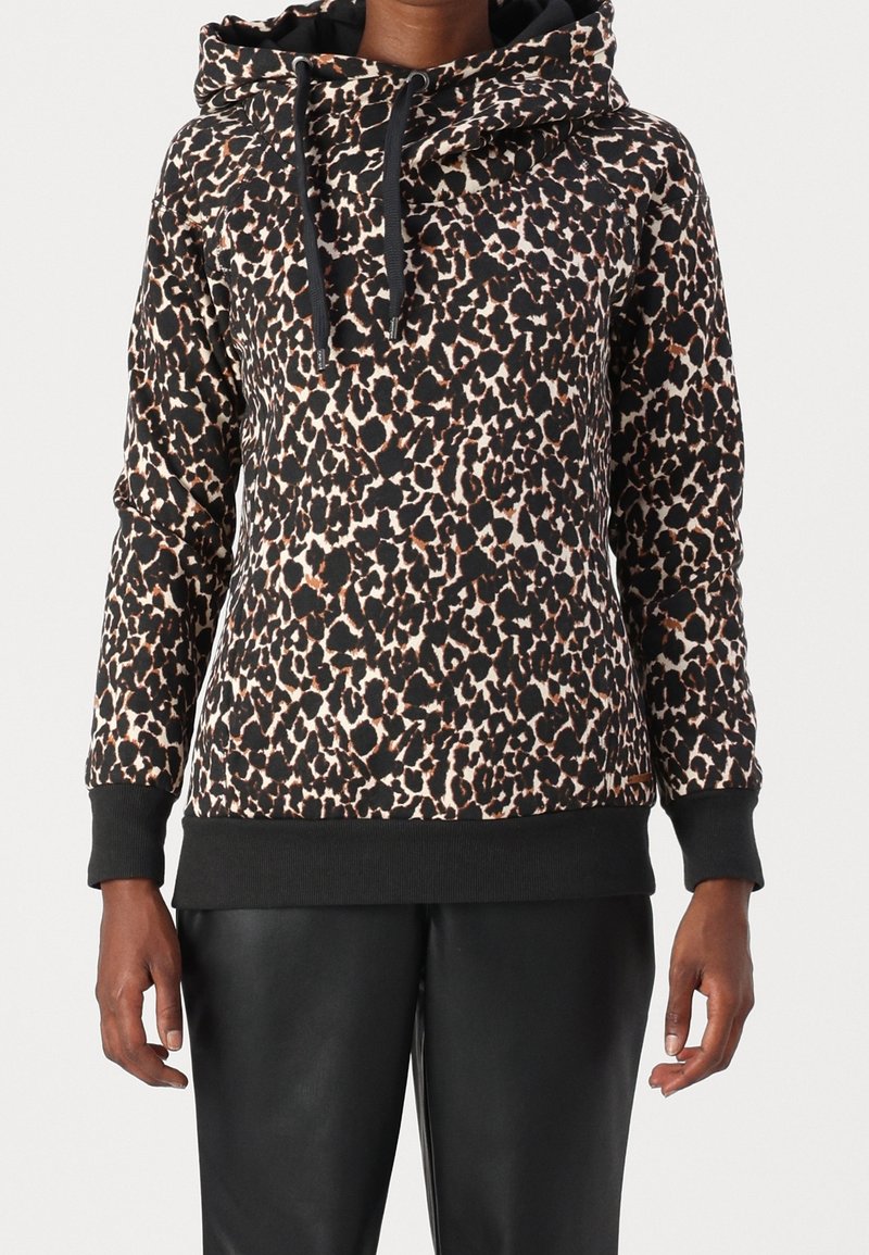 Personne portant un sweat à capuche imprimé léopard noir et beige avec des cordons noirs et un pantalon noir, posant devant un fond uni.