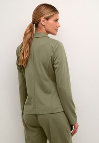 Olivinės spalvos, prigludusi striukė, pagaminta iš minkštos medžiagos, su apykakle, atgaliniu siūlės detalizavimu ir ilgomis rankovėmis, suderinta su atitinkamais džinsais.