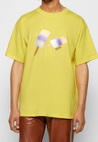 Camiseta de algodón amarilla con un gráfico de dos polos en púrpura y blanco, con un logo de MCM. Corte relajado, mangas cortas.