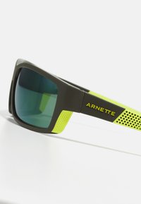 Arnette solbriller med mørkebrun ramme, grønt glass og lysegule detaljer med perforert design på sidene.