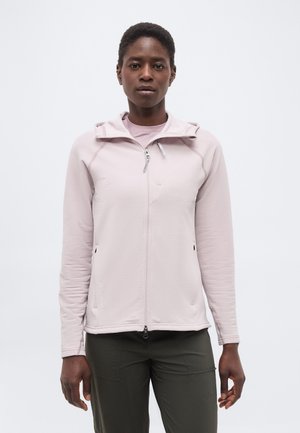 Joven mujer negra con chaqueta rosa claro con cremallera y pantalones oscuros, de pie contra un fondo blanco liso, mirando hacia adelante.