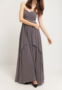 Žena ve strapless šedé šifónové večerní šaty s nařaseným korzetem a zdobeným pasem, stojící s jednou ohnutou paží a druhou paží natáhnutou.