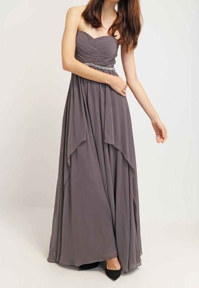 Žena ve strapless šedé šifónové večerní šaty s nařaseným korzetem a zdobeným pasem, stojící s jednou ohnutou paží a druhou paží natáhnutou.