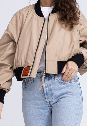 Personne portant une veste bomber beige avec col et poignets noirs, chemise blanche et jean taille haute bleu clair, main reposant près de la boucle de ceinture.