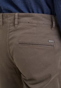 Bruna bomullsbukser med en bakficka med horisontell öppning, metalldekal och en mjuk matt yta.