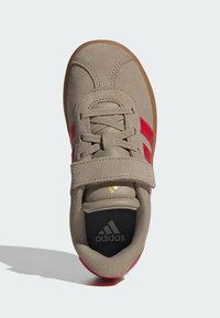 Hnedé semišové topánky s červenými pruhmi a suchým zipsom. Majú polstrovanú stielku a gumovú podrážku v béžovej farbe. Logo je viditeľné na jazyku.