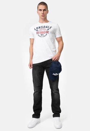 Noor mees kannab valget Lonsdale graafilise trükiga t-särki, musti teksapükse, valgeid tossusid, hoiab paremas käes tumel sinist Lonsdale mütsi.