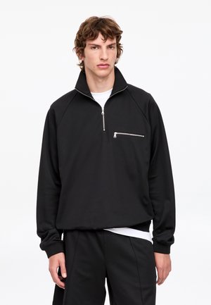 Giovane uomo in piedi, che indossa un pullover nero con mezza zip e tasca con zip sul petto, sopra una camicia bianca e pantaloni neri, su uno sfondo semplice.