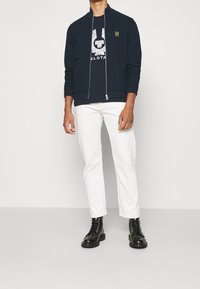 Mörkblå zip-up jacka med högt krage, svart grafisk t-shirt under, vita byxor och svarta snörkängor. Rent linjer och avslappnad passform.