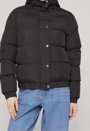 Person iført en sort dunjakke med trykknapper og elastiske manchetter, kombineret med løstsiddende lyseblå jeans.