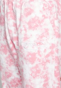 Calças de sudoeste cor-de-rosa claro com um padrão marmoreado, faixa elástica na cintura e bolsos laterais. Apresenta um logotipo branco em relevo na perna esquerda.