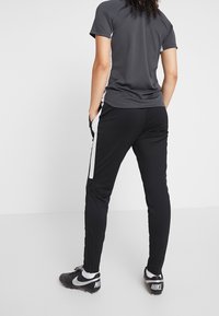 Person steht mit Händen in den Taschen, trägt Schwarz-Weiß-Trainingshosen, ein dunkelgraues Kurzarmshirt und schwarze Nike-Stollenschuhe auf weißem Boden.