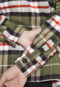 Zielona koszula w kratę z flaneli, z czerwonymi, czarnymi i białymi kratkami; miękka w dotyku; detale przy mankietach z guzikami; podwinięte rękawy, odsłaniające ręce.