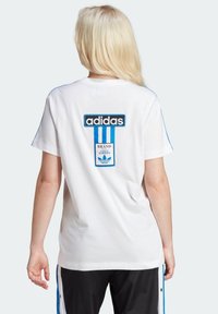 Vit bomullströja med svart och blå Adidas-logotyp och grafik på baksidan, med blå accentlinjer längs axelsömmarna.