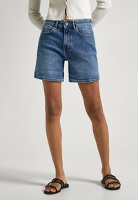Pantalones cortos de denim en azul claro, con una cintura de altura media, ajuste relajado y dobladillos enrollados, combinados con sandalias negras y un cárdigan blanco.