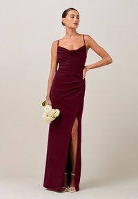 Frau in einem bordeauxfarbenen, ärmellosen Abendkleid mit Beinschlitz, die weiße Blumen hält, weiße High Heels und goldenen Schmuck trägt, vor neutralem Hintergrund.
