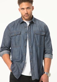 Donkerblauwe denim shirt met knoopsluiting aan de voorkant, twee borstzakken, opgerolde mouwen, contrasterende stiksels en een versleten textuur, gecombineerd met een wit T-shirt.