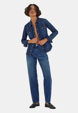 Giovane persona che indossa una camicia di jeans blu scuro e jeans, in piedi con le mani vicino ai bottoni della camicia, indossa mocassini neri.