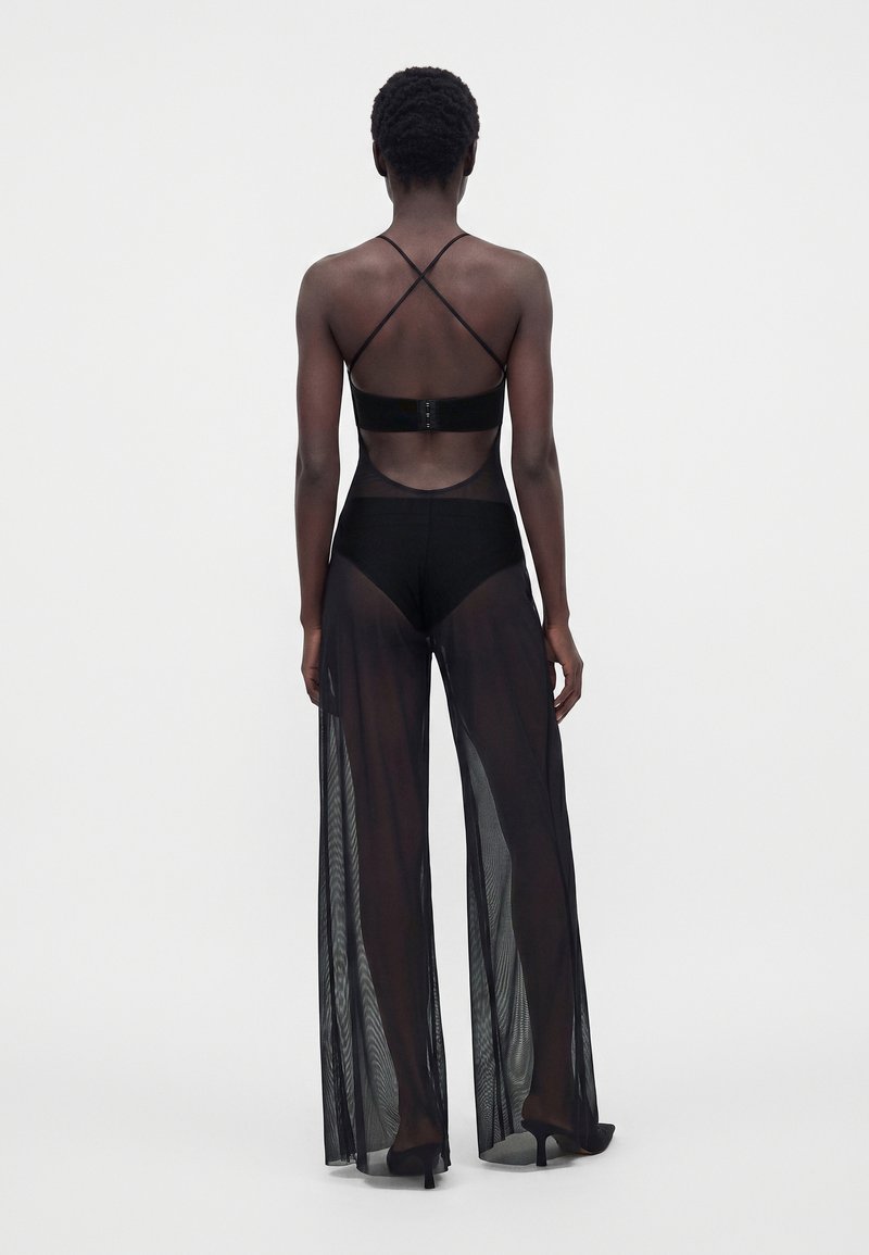 Femme portant un pantalon noir large et transparent et un haut noir avec des fines bretelles croisées, vue de dos sur un fond uni.