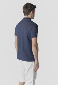 Uomo di spalle che indossa una polo blu navy a maniche corte con rifiniture rosse e pantaloncini cargo bianchi, su uno sfondo semplice.
