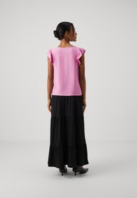 Top rosa con texture e maniche a volant, abbinato a una gonna maxi nera a strati. La modella indossa tacchi a punta neri, vista da dietro su uno sfondo semplice.