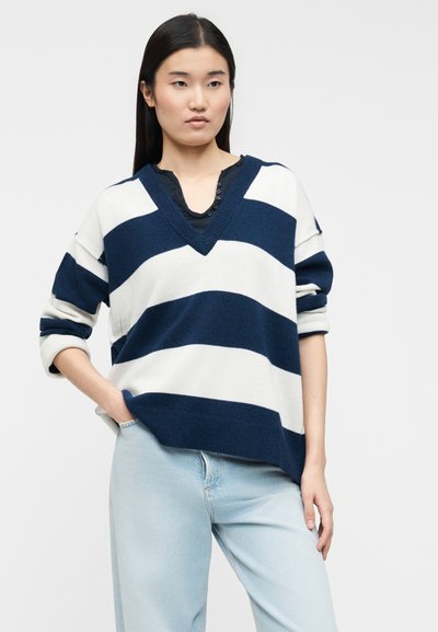 Νεαρή γυναίκα που φοράει μπλε ναυτικό και λευκό ριγωτό πουλόβερ με V λαιμόκοψη και μανίκια που έχουν γυριστεί, καθώς και ανοιχτόχρωμο τζιν, στέκεται με το ένα χέρι στην τσέπη.