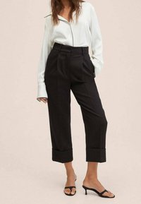 Sorte højtaljede bukser med plisseret design og opsmøg på nederste kant, parret med en hvid satin langærmet skjorte og sorte hælede sandaler.