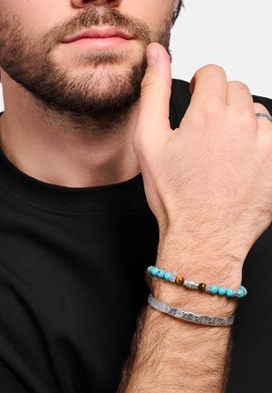 Uomo con la barba che tiene la mano vicino al volto, indossa una camicia nera, un braccialetto di perline turchesi e un braccialetto argentato decorato.