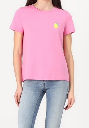 Kobieta ubrana w różowy t-shirt z krótkim rękawem z małą żółtą grafiką cytryny na lewej piersi, w połączeniu z jasnoniebieskimi dżinsami.