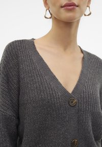 Harmaa neulottu neuletakki, jossa V-aukko ja kolme suurta nappia sekä teksturoidun näköinen kangas. Malli käyttää suuria kultaisia rengaskorvakoruja.