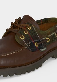 Chaussure en cuir marron avec col en tissu tartan vert, lacets en cuir, œillets métalliques jaunes et semelle épaisse en caoutchouc avec étiquette Barbour.