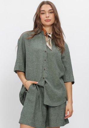 Vrouw met lang bruin haar, die een losse groene blouse met knopen draagt, bijpassende korte broek en een patroon halsdoek, staand tegen een effen achtergrond.