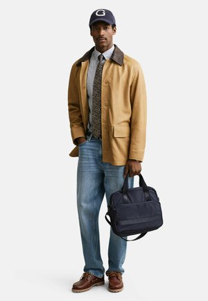 Mann trägt eine beige Jacke, gemusterten Krawatte, Jeans, braune Schuhe, marineblauen Hut mit „G“ und hält eine marineblaue Aktentasche an den Griffen.