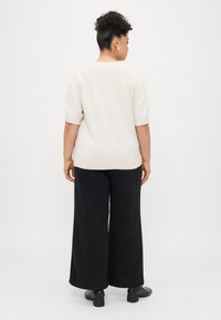 Ανοιχτό μπεζ κοντομάνικο πουλόβερ και μαύρα παντελόνια με φαρδιά ποδιά. Το πουλόβερ έχει χαλαρή εφαρμογή, ενώ τα παντελόνια έχουν λεία υφή με ελαφριά γυαλάδα.